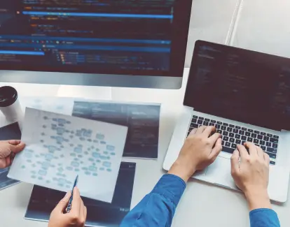   Un ordinateur de bureau et un ordinateur portable placés devant deux bras d'individus en signe de travail ou de collaboration.